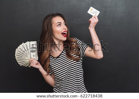 Picture of smiling young lady standing over grey wall and holding money and debit card in hands. Looking aside.
