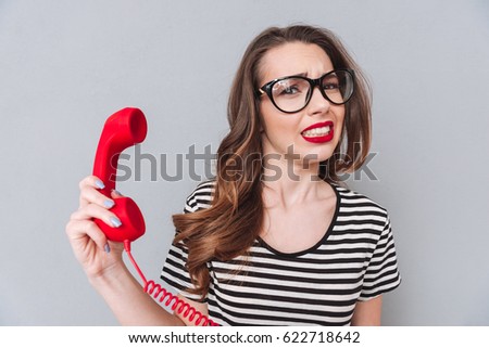 Image of confused young lady standing over grey wall holding phone. Looking at camera.