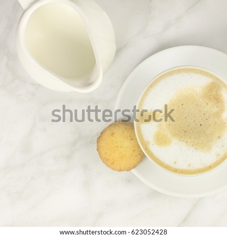 A square photo of a cup of coffee with milk with a butter cookie, and milk jug, shot from above on a light texture for copy space