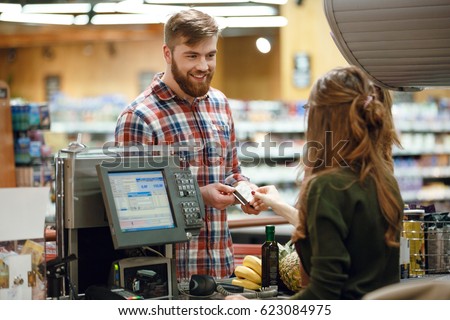 Photo of happy young man standing in supermarket shop near cashier's desk holding credit card. Looking aside.