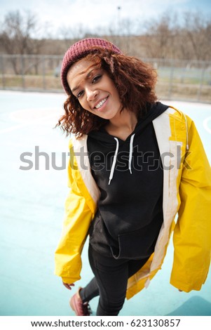 Picture of happy african curly young woman wearing yellow coat walking outdoors. Looking at camera.