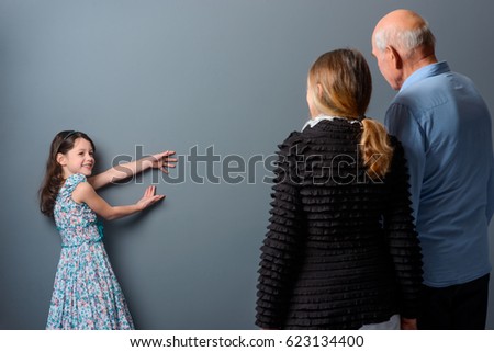 Girl shows something abstract on the wall. Grandparents staying turned their backs to the camera. Place for text or picture in the middle of the photo, gray background.
