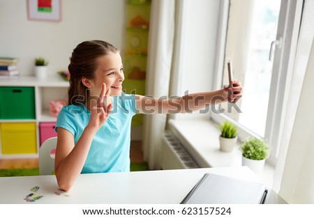 people, children and technology concept - girl with laptop computer and smartphone taking selfie at home and showing peace hand sign