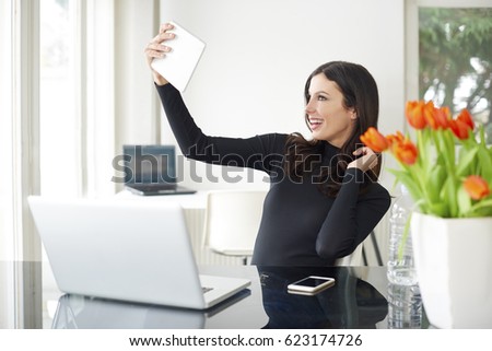 Shot of a young woman using digital tablet and taking selfie while sitting at office desk in front of laptop.