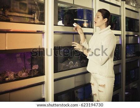 Cheerful teenager in aquarium shop points to interesting colored fish on several rows of shelves with aquariums
