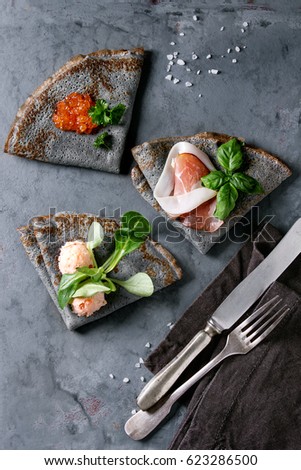 Black pancakes crepes with variety of salt filling red caviar, ham, fish salmon pate, green salad with vintage cutlery over gray texture metal background. Top view