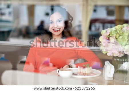 Woman drinking hot cappuccino coffee and eating cake at cafe. Photo taken through the window