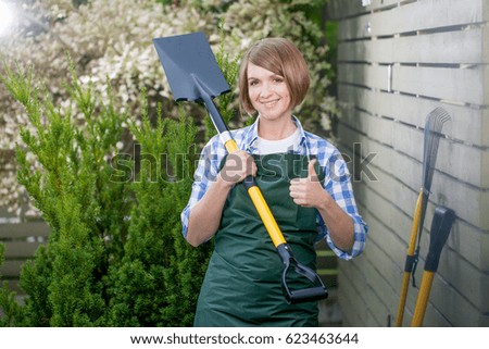 smiling young woman professional gardener or florist standing in the yard with shovel and showing thumb up sing. garden worker posing outdoors. gardening service and planting concept
