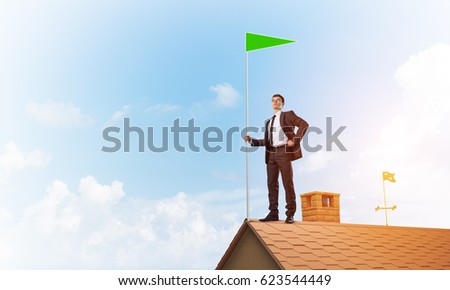 Businessman standing on house roof and holding green flag. Mixed media