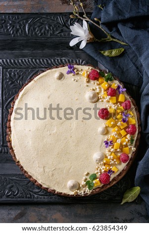 Homemade chocolate tart decorated by mango, raspberries, mint, puffed rice and edible flowers served with blue textile and magnolia over dark ornate metal background. Top view. Comfort food concept.