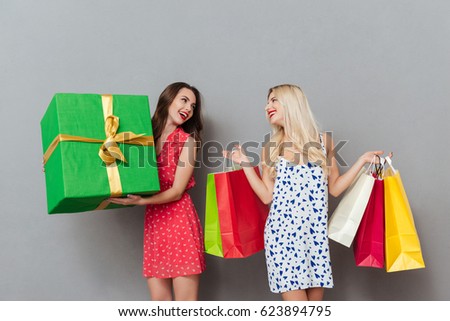 Photo of amazing young two ladies friends with bright makeup lips standing over grey wall and posing with shopping bags and gift. Looking aside.