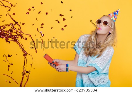 portrait of funny blond woman in birthday hat and red confetti on yellow background. Celebration and party.