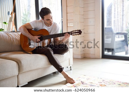 Pretty young man playing guitar while sitting on sofa in light living room Royalty-Free Stock Photo #624050966