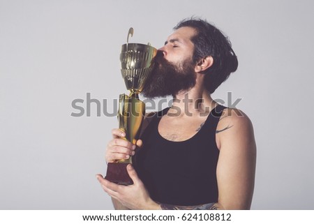 bearded man, long beard, brutal caucasian hipster with moustache in black vest kissing gold champion cup with happy face on grey background, unshaven guy