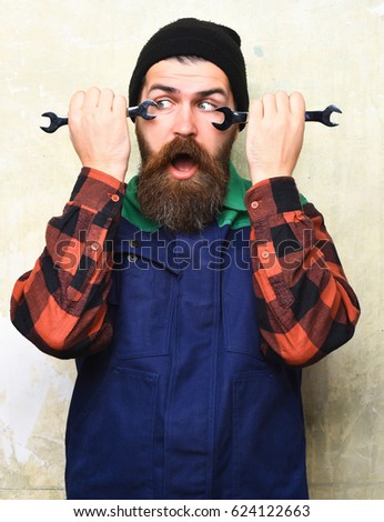 Bearded man, long beard. Brutal caucasian mechanic, surprised unshaven hipster in black hat, checkered shirt and uniform holding metallic wrenches on texture studio background