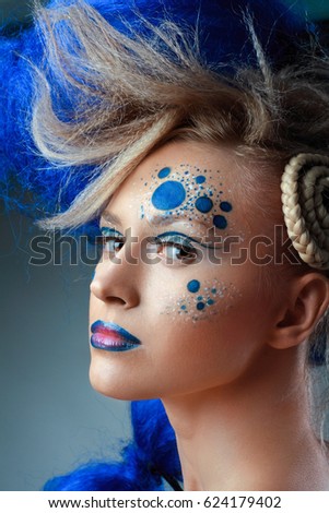 close up portrait of young beautiful woman wearing artistic make up and blue wig posing next to color background