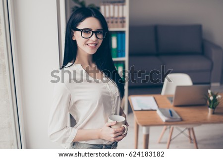 Photo of pretty young beautiful woman stands edgeways in the office near the window, holds mug of tea  in her hands and looks directly into the camera