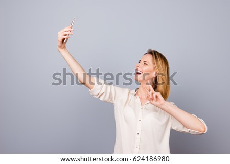Photo of pretty young happy laughing woman taking selfie using her mobilephone, two-fingers gesturing