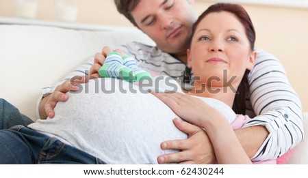 Portrait of a pregnant woman holding baby shoes and of her husband lying on the sofa at home
