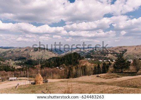 Mountain landscape with wooden houses, a haystack and snow on the tops of the mountains. Background of spring mountains. Clouds, sky and air. Recreation and tourism. Ukrainian Carpathians