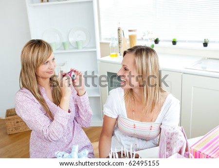Two glowing female friends taking pictures during a birthday party in the kitchen at home