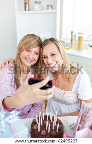 Two radiant female friends taking pictures during a birthday party in the kitchen at home