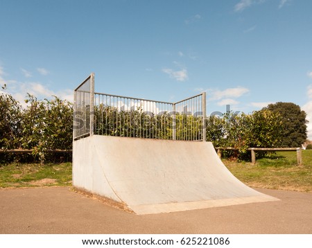 An Empty and Unused Ramp Half Pipe at the Skate Park in the Country Park in the UK Shining in the Sunlight of the Day and with A Shrub Hedge Behind the Bars Protecting it Fenced in the Spring