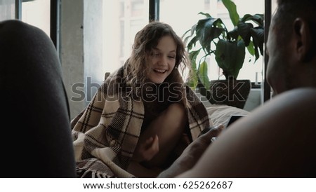 Man takes photos on film camera of woman. Female laughing, wrapping in blanket. Multiethnic couple looks happy together.