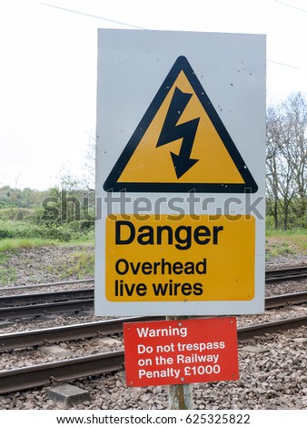 A Railway Safety Sign Saying Danger Overhead Live Wire Warning Do Not Trespass On the Railway Penalty a thousand pound in Black, Yellow, Red, and white with Track in Background in Spring in UK