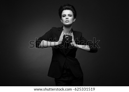 Young pretty woman with short dark hair wearing black blazer posing in dark studio, holding old photo camera in her hands, in black and white