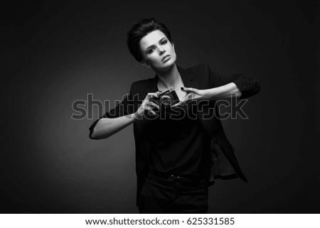 Young serious beautiful woman with short dark hair wearing black blazer posing in dark studio, holding old photo camera in her hands, in black and white