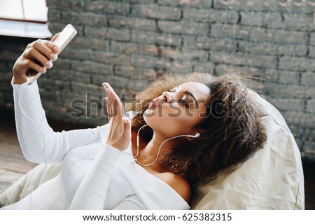 Happy modern African American young girl talking on video chat and lying at home with loft interior, communication concept. Smiling hipster girl making self portrait on her telephone at home