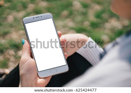 Girl with smartphone in hands with mock up white screen of blank screen sits in park on open space, screen for content integration. Hands holding gadget on blurred backdrop, 