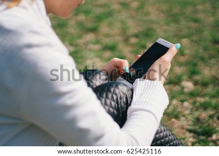 Beautiful young European girl sits on the grass in the park and uses a smartphone, concepts of using gadgets in a natural environment