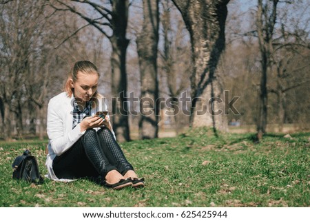 Beautiful young European girl sits on the grass in the park and uses a smartphone, concepts of using gadgets in a natural environment