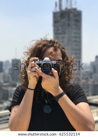 Portrait of a long curly haired man taking a photo with an analogical camera, blurry buildings at background