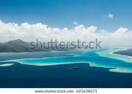 Aerial view on lagoon of Raiatea island in French Polynesia with blue and turquoise water, barrier reef, blue sky, hills with tropical forest and white clouds 