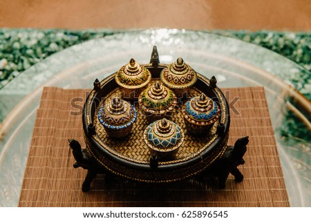Traditional Thai famous porcelain benjarong ceramic decorative bowls on wicker plate with elephants standing on rattan table with glassy surface with abstract falling water wall background. Ceremony.