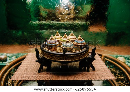 Traditional Thai famous porcelain benjarong ceramic decorative bowls on wicker plate with elephants standing on rattan table with glassy surface with abstract falling water wall background. Ceremony.