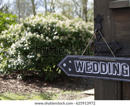 Wedding this way sign in black with white text hanging on ornate iron door handle with summer garden in the background