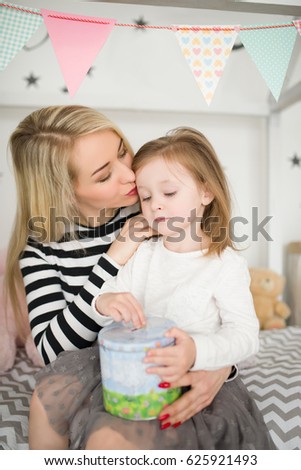 Attractive mother kisses her a little daughter who sits in mother's lap and keeps moneybox in hands, in the room