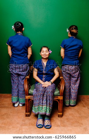 Photo from back of two Thai ethnic women pissing to wall in similar traditional exotic spa costumes and third sitting in rattan chair and looking at camera on green background. Psychological fashion.