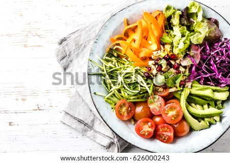 Vegan buddha bowl. Bowl with fresh raw vegetables - cabbage, carrot, zucchini, lettuce, watercress salad, tomatoes cherry and avocado, nuts and pomegranate. White background, top view, copy spae