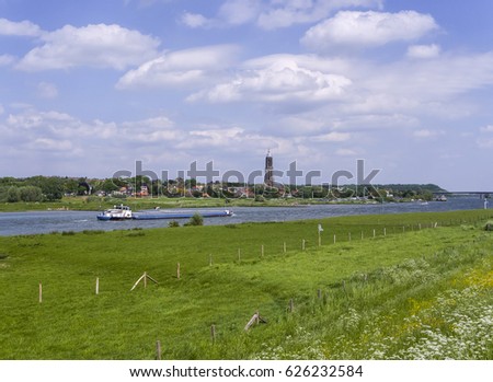 Small town near the river with high church tower, a ship sailing past and grasslands with flowers in spring