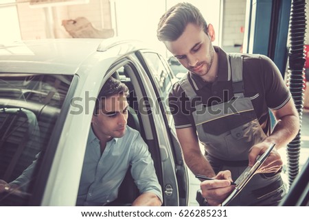 At the auto service. Handsome young auto mechanic in uniform is talking with a client and showing a document to fill