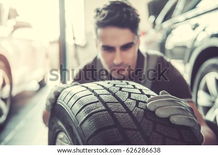 Handsome young auto mechanic in uniform is examining a tire while working in auto service Royalty-Free Stock Photo #626286638