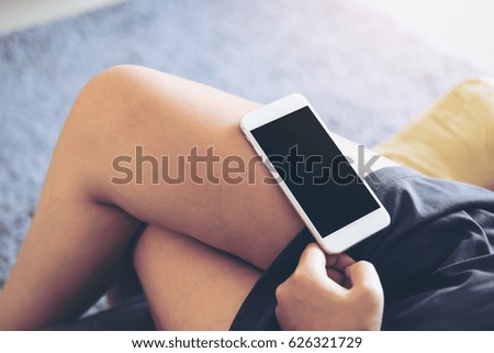 Mockup image of white mobile phone with blank black screen on woman's thigh with gray rug background