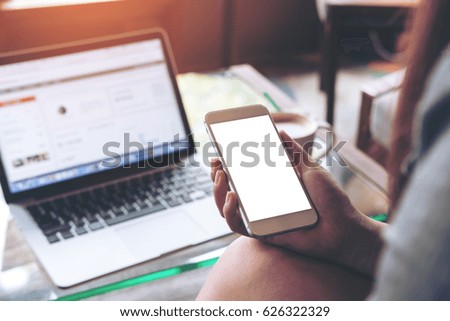 Mockup image of a woman holding mobile phone with blank white screen while using laptop with coffee cup on the table in office
