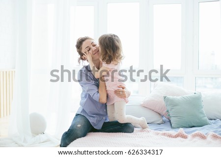 Young mother and her little daughter hugging and kissing on bed on a sunny morning. Soft pastel colors, pink and blue. Selective focus. Love and care. Happy family time on weekend. Mother's Day