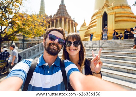Successful traveling couple in love taking a selfie on phone  at Grand Palace temples in Bangkok  Pretty girl and her handsome boyfriend with beard having fun, crazy emotional faces , laughing.
